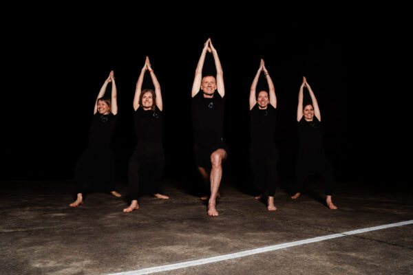 Un groupe de personnes vêtues de sombre montre de manière synchrone une position de yoga sur un fond sombre.